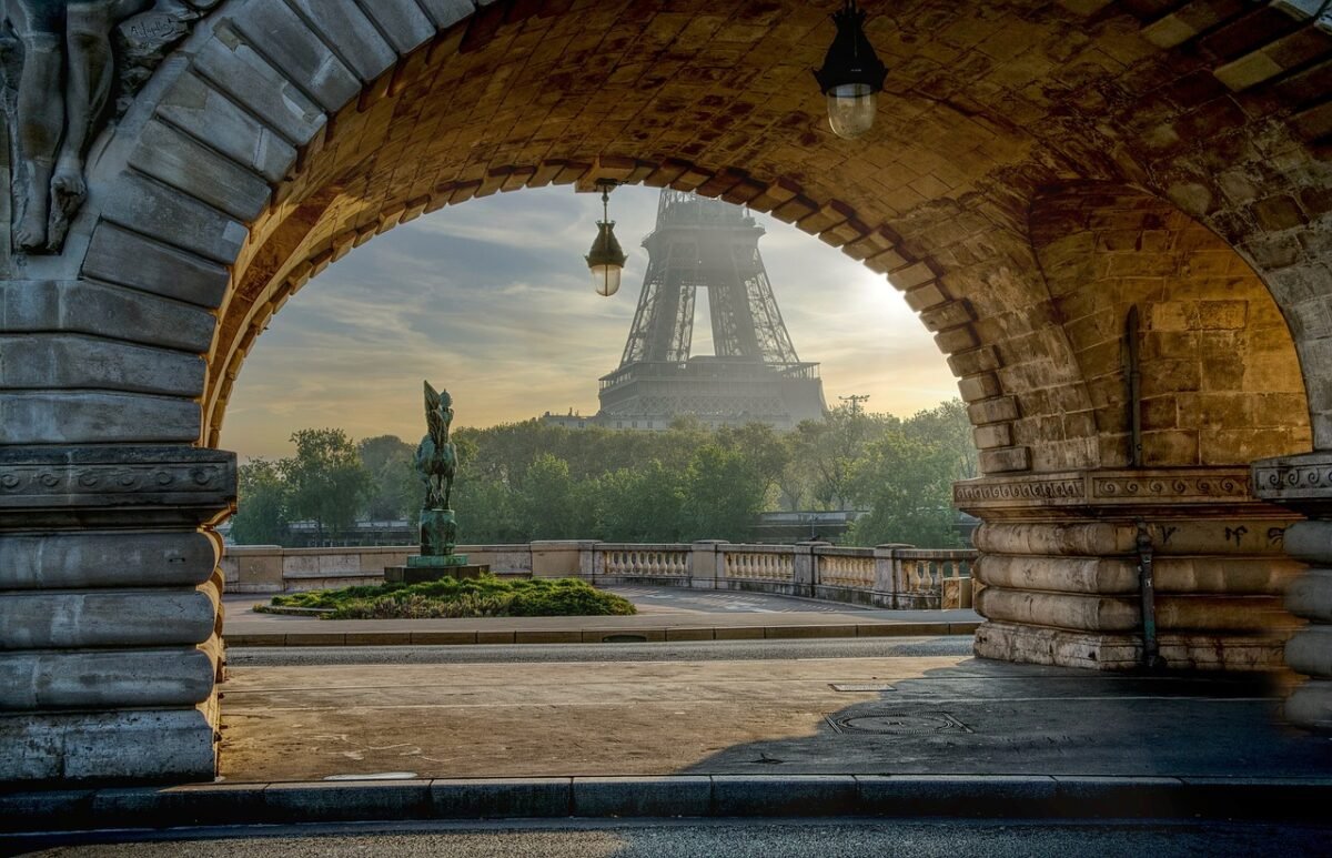 View of the Eiffel Tower at sunrise, framed beautifully through the stone arch of the Pont de Bir-Hakeim bridge in Paris. The scene captures the tranquil morning light, lush greenery, and an elegant sculpture in the foreground, highlighting the romantic charm of the city.