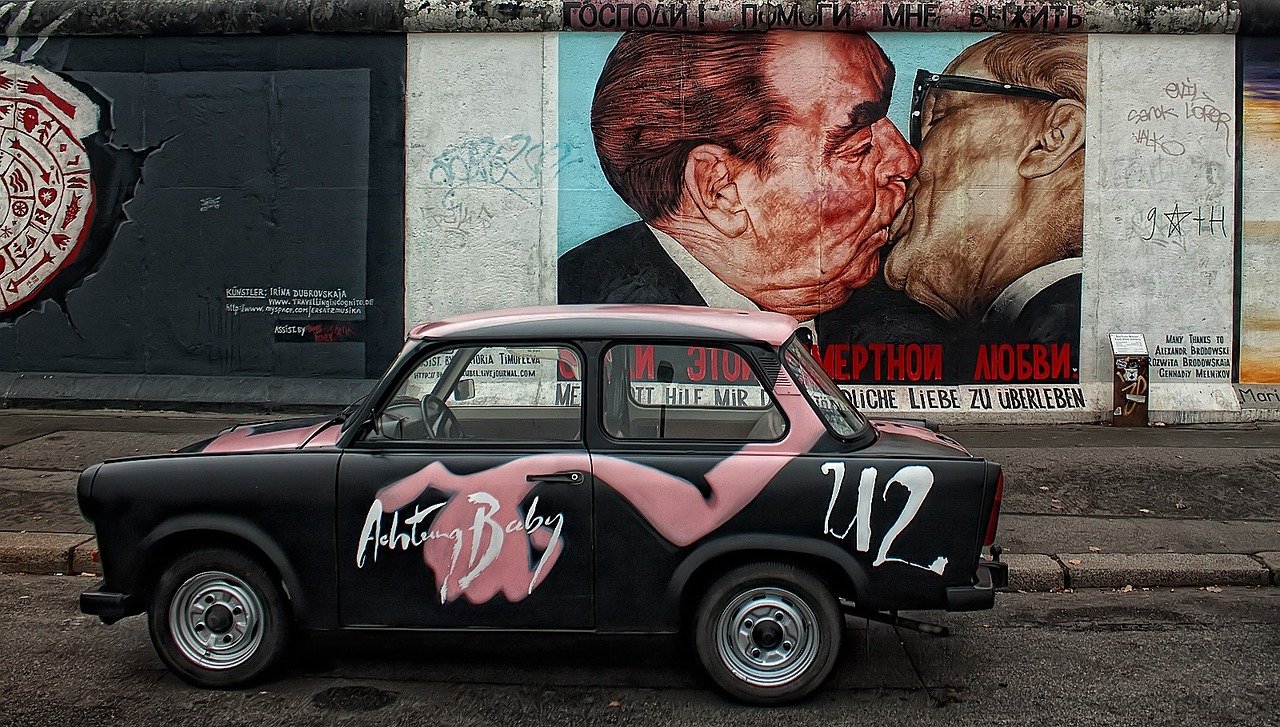 Iconic scene at the East Side Gallery in Berlin featuring the famous mural 'The Fraternal Kiss,' a symbolic piece of street art on a preserved section of the Berlin Wall. In front of the mural is a classic Trabant car painted with vibrant designs, embodying the fusion of historical significance and artistic expression in Berlin's cultural landscape.
