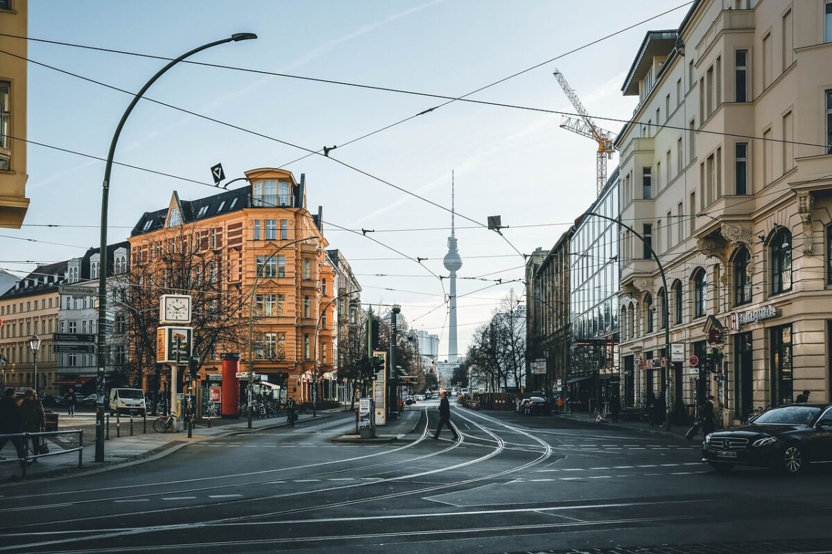 Street view of Berlin featuring the iconic Fernsehturm (TV Tower) in the background, surrounded by a blend of modern and classic architecture, with a vibrant orange building in the foreground. The scene includes tram lines, a clock, and people crossing the street, capturing the city's dynamic urban atmosphere.