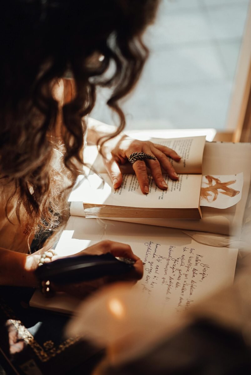 A close-up of a person with curly hair writing in a notebook while reading a book, with sunlight streaming in, creating a warm and contemplative atmosphere.