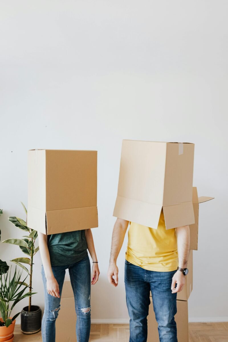 A playful image of a couple standing in a room with moving boxes on their heads, symbolizing the fun and challenges of moving, ideal for real estate marketing or homeownership campaigns.