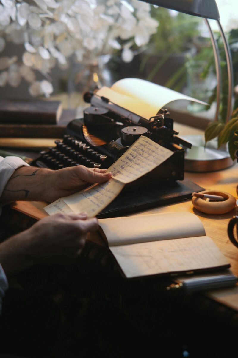 A cozy workspace featuring a vintage typewriter, a lamp casting warm light, and a person holding a handwritten note while sitting at a wooden desk surrounded by plants and books.