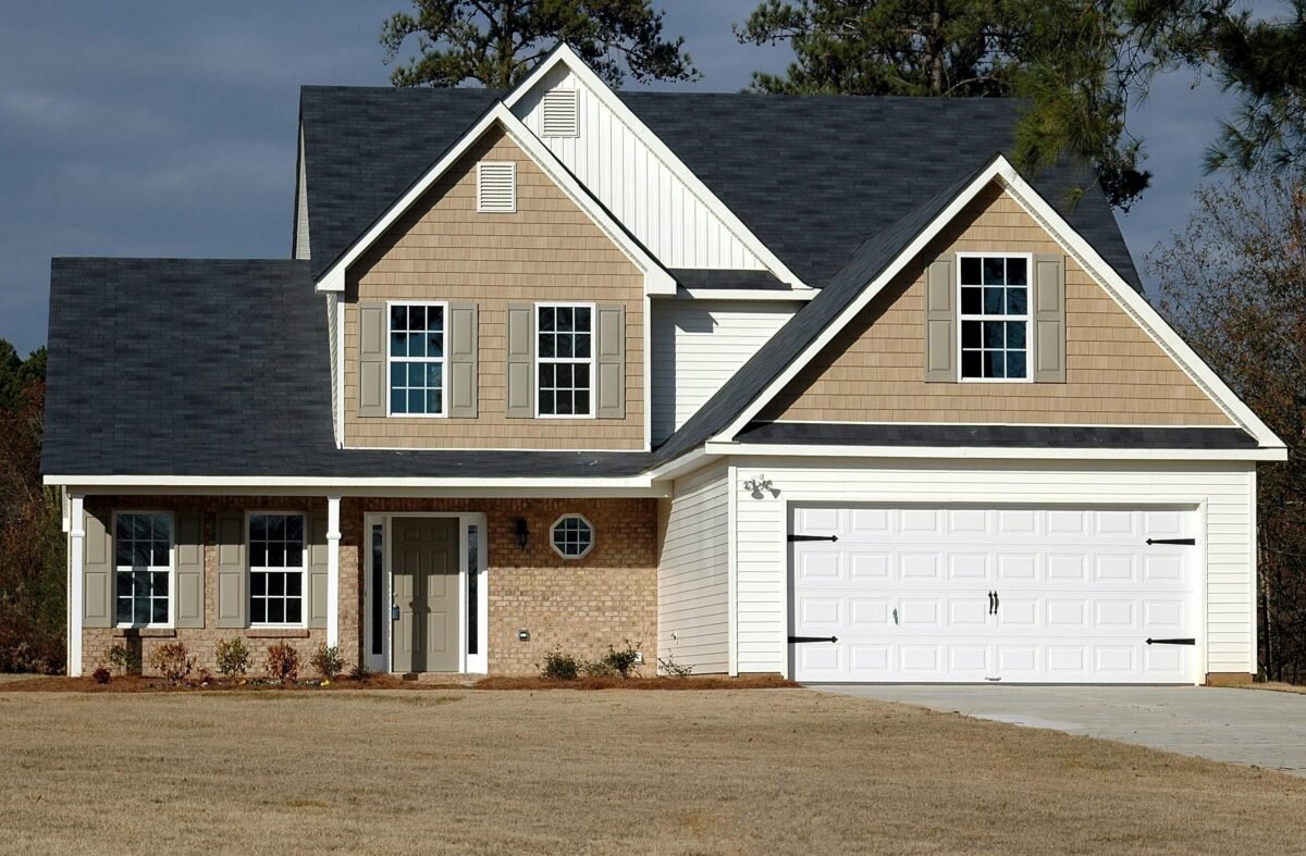 A modern suburban house with a beige brick facade, white garage doors, and a well-maintained front yard.