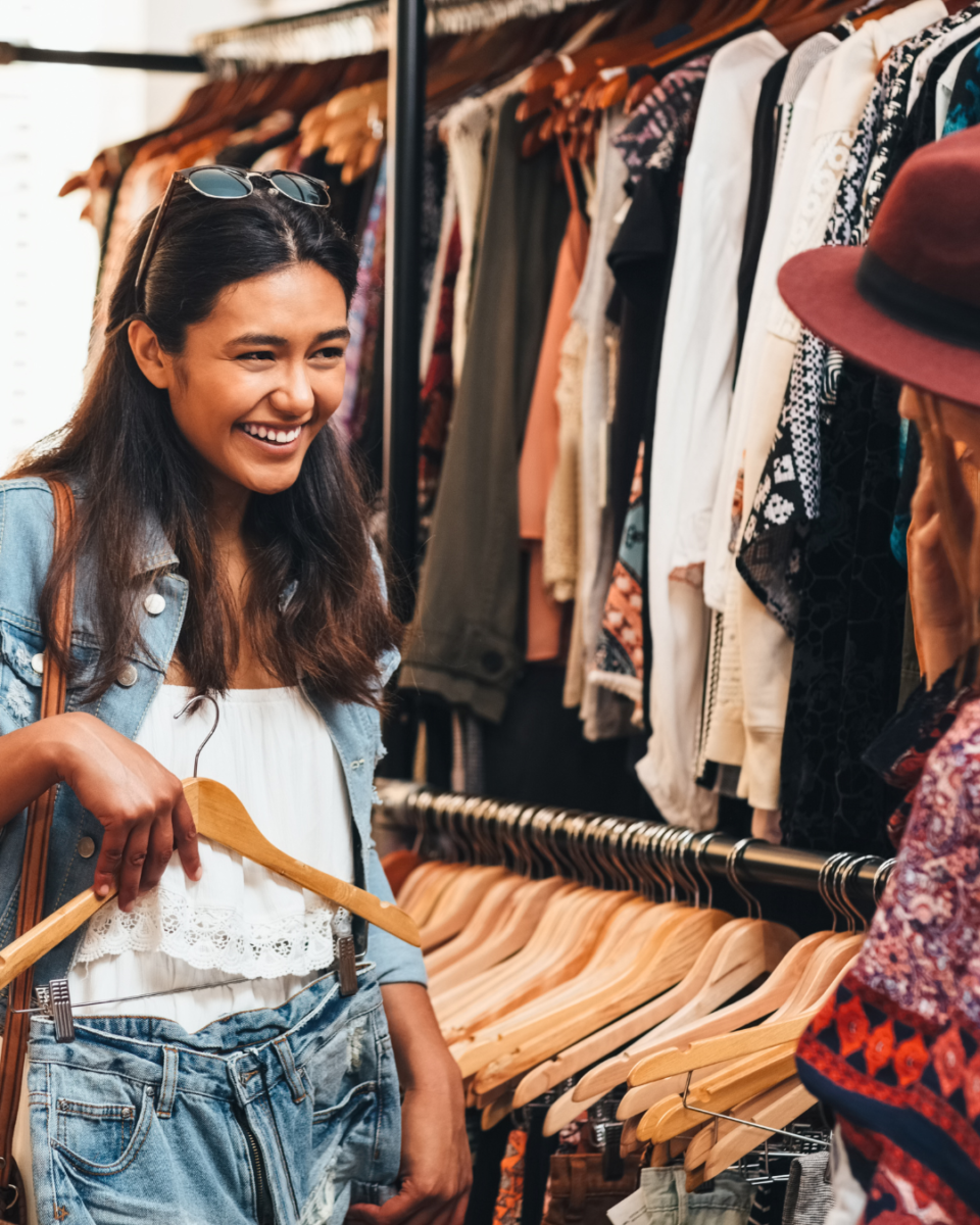A young woman shopping in a boutique, smiling while holding a clothing hanger. She is engaging in conversation with another shopper. The target audience for this image includes fashion-conscious young adults, boutique shoppers, and those interested in trendy, personalized shopping experiences.