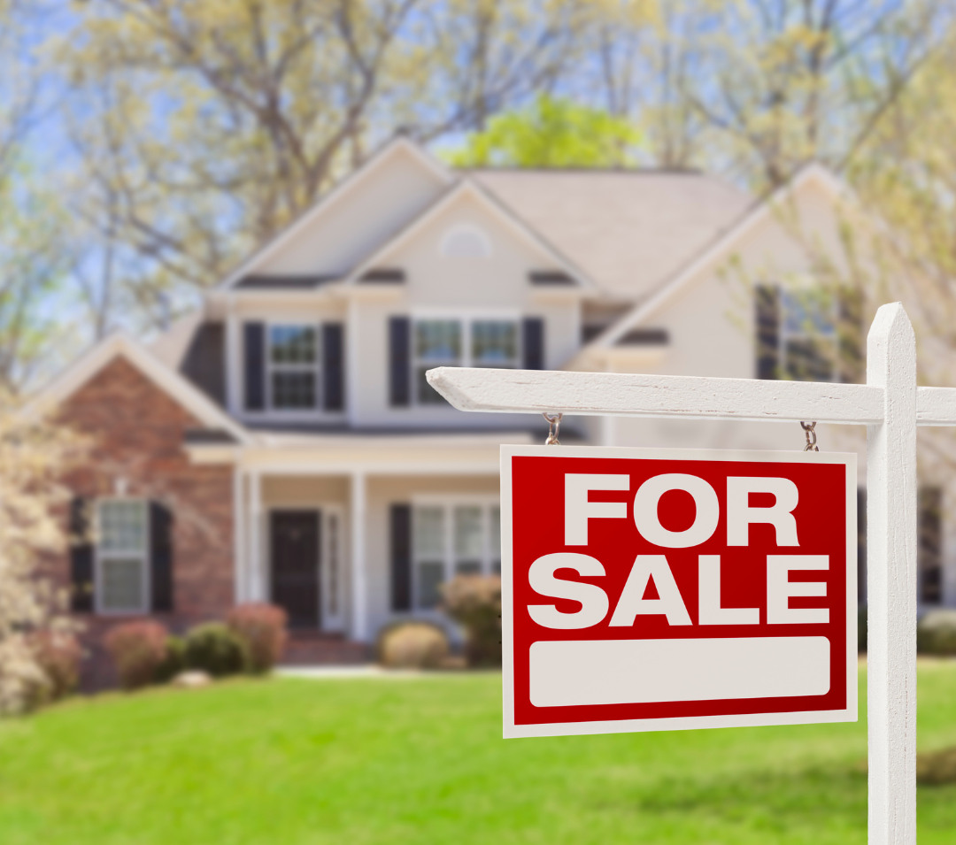 A red and white "For Sale" sign hanging from a white post in front of a suburban house with a green lawn and trees in the background.