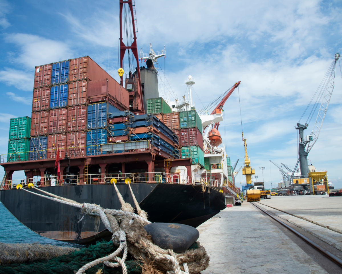 A cargo ship loaded with colorful shipping containers docked at a busy port, with cranes and trucks in the background. This image represents global trade and the challenges businesses face as they navigate the tariff effect.